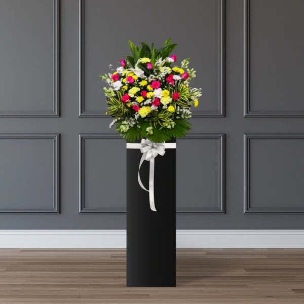 Flower arrangement with white lilies, pink roses, yellow and white carnations, yellow mums, and white matthiola on a black stand with a silver bow.