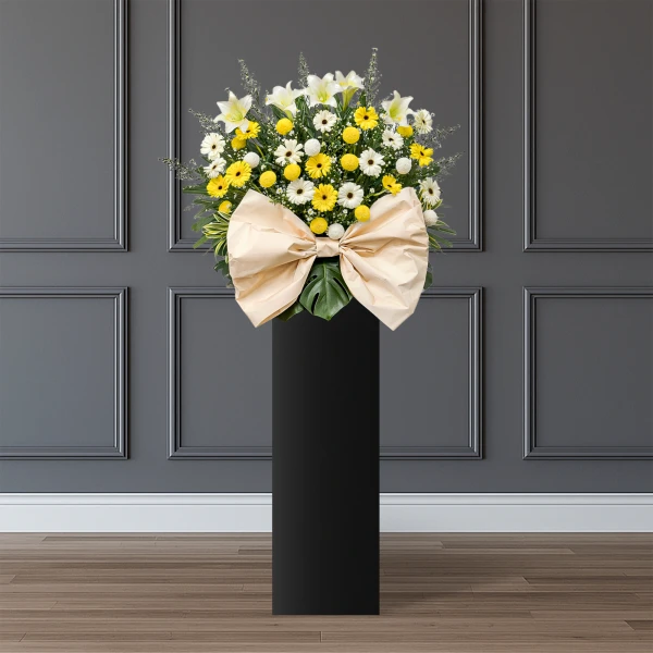 A large floral arrangement of white lilies and yellow daisies with a beige bow, atop a black pedestal.