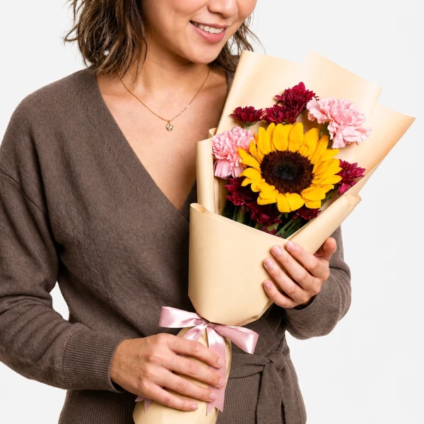 Woman holding a bouquet with a sunflower, pink carnations, and red chrysanthemums, wrapped in light brown paper.