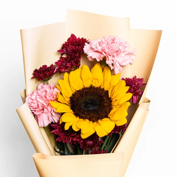A sunflower bouquet with pink carnations and dark red chrysanthemums, wrapped in tan paper.
