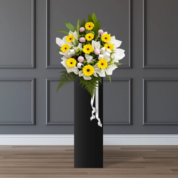 Yellow gerberas and purple pingpong mums in a white bouquet on a black stand with white ribbon.
