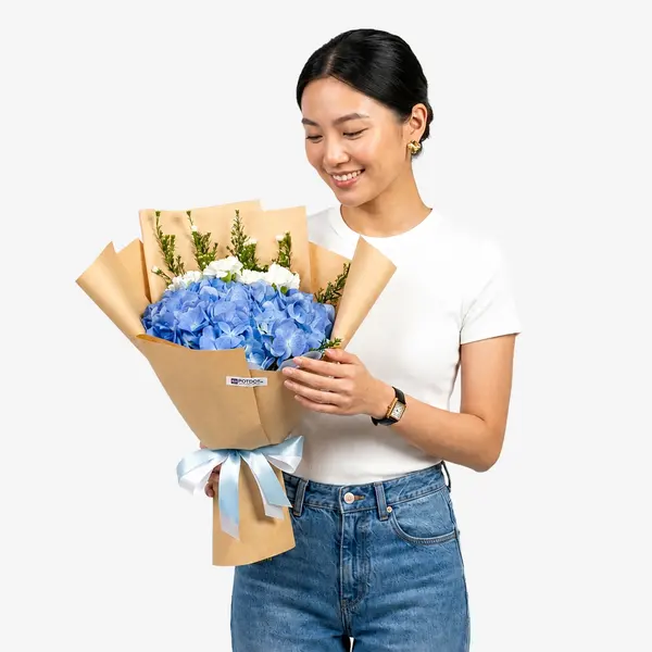 Person holding a bouquet of blue hydrangeas, white carnations, and asters wrapped in brown paper with a blue and white ribbon.