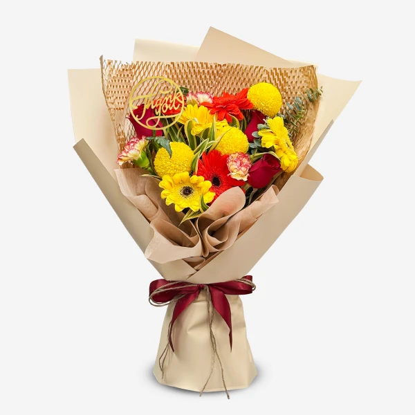 Colorful bouquet of red roses, yellow gerberas, and red carnations with a "Grateful" cake topper, wrapped in brown paper and tied with a maroon ribbon.