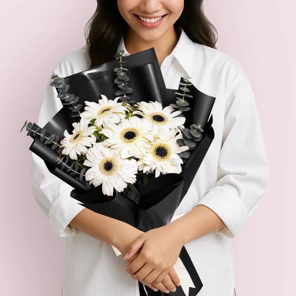 Woman in a white shirt holding a bouquet of white gerberas and eucalyptus wrapped in black paper.