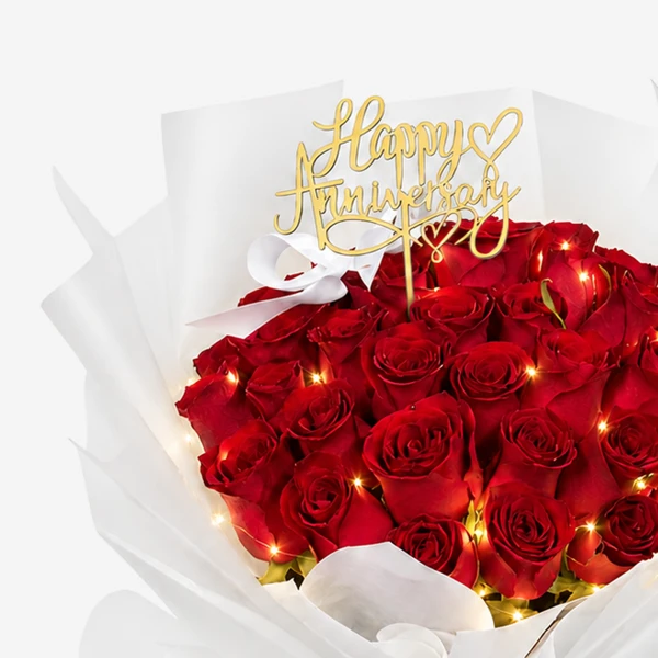 A bouquet of red roses with a "Happy Anniversary" cake topper and fairy lights.