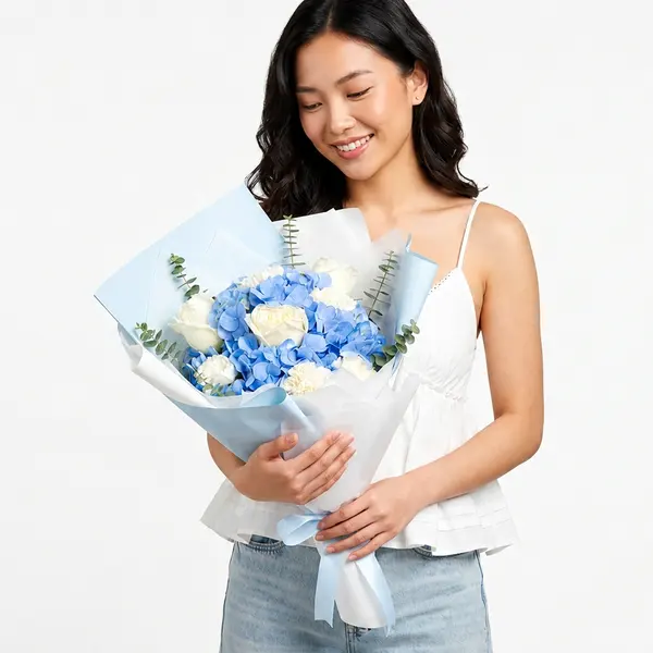 A person holds a bouquet of blue hydrangeas, white roses, and carnations, wrapped in blue and white paper.