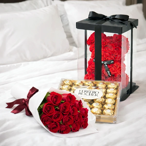 Red rose bouquet, Ferrero Rocher chocolates, and a red rose bear in a clear box on a white bed.