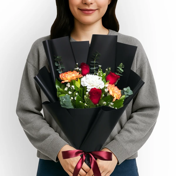 A person holds a bouquet of red roses, orange and white carnations, and greenery wrapped in black paper, tied with a red ribbon.