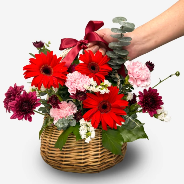 Red gerberas, pink carnations, and violet chrysanthemums in a woven basket with a red ribbon.