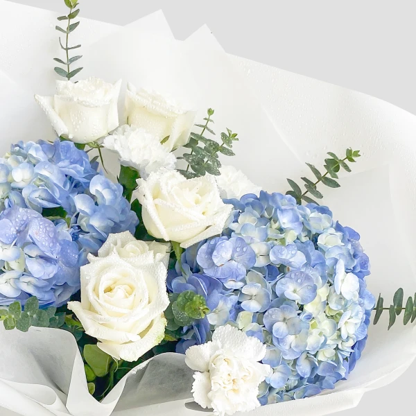 White roses, blue hydrangeas, and white carnations with eucalyptus in white paper wrapping, water droplets visible.