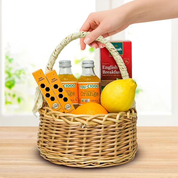 Wicker basket with orange juice, lemon, orange, tea box, and mini straws, held by a hand.