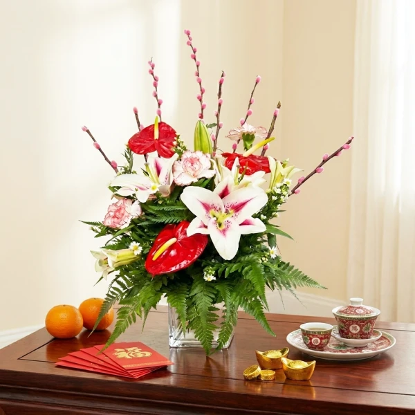 Floral arrangement with lilies, anthuriums, and pussy willow branches on a wooden table with oranges and red envelopes.