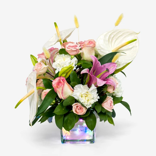 A floral arrangement in a clear cube vase with fairy lights, featuring pink roses, white lilies, and white calla lilies.