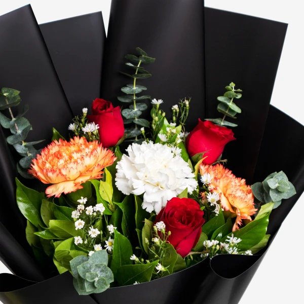 A vibrant bouquet of red roses, orange carnations, and white blooms with green foliage, wrapped in black paper.