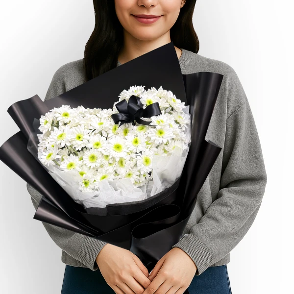 Heart-shaped bouquet of white chrysanthemums with a black ribbon, held by a person in a gray sweater.