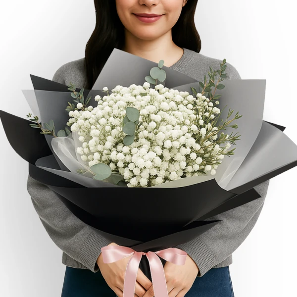 A person holds a large bouquet of white baby's breath flowers wrapped in black and grey paper, tied with a pink ribbon.