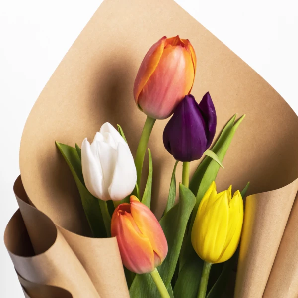 A bouquet of five colorful tulips (white, purple, yellow, and two red-orange) wrapped in brown paper.