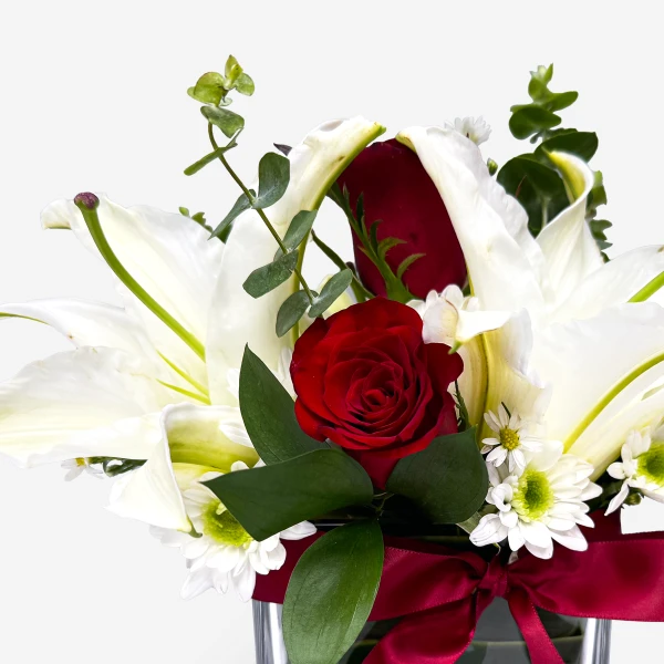 Red roses and white lilies in a clear vase with a red ribbon and white chrysanthemums.