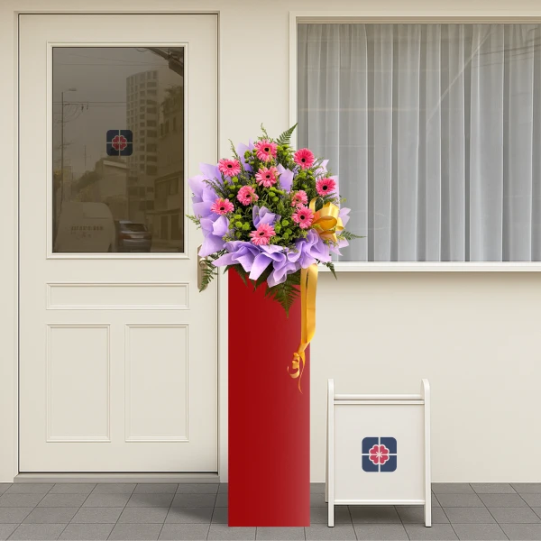 A grand opening flower arrangement of pink gerberas on a red stand, with a white sign.