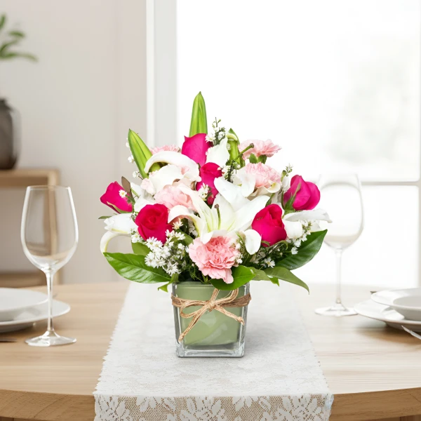 A square glass vase holds a vibrant floral arrangement of pink roses, white lilies, and pink carnations.