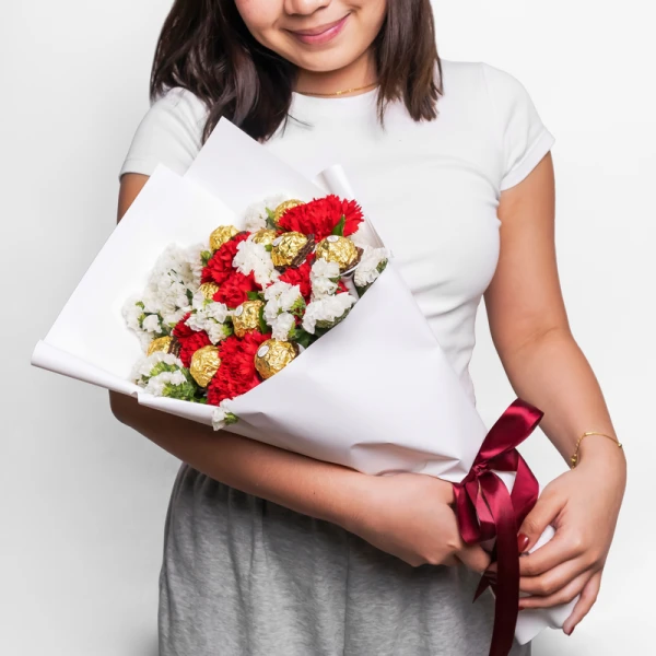 Woman holding a bouquet of red carnations, white statice, and Ferrero Rocher chocolates, tied with a burgundy ribbon.