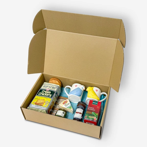 Open gift box filled with crackers, cheese, jam, tea, and two novelty mugs.