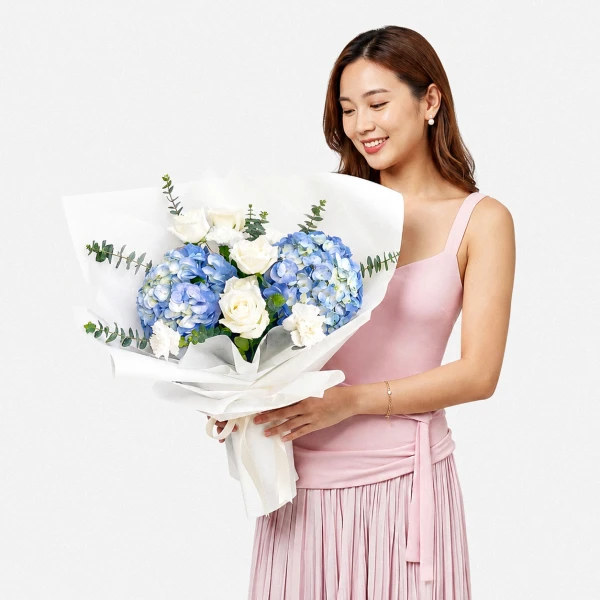 A woman holds a bouquet of blue hydrangeas, white roses, and white carnations, elegantly wrapped in white paper.