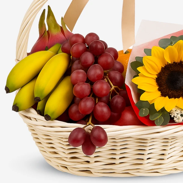 Wicker basket with bananas, dragonfruit, grapes, orange, apple, and a sunflower bouquet.