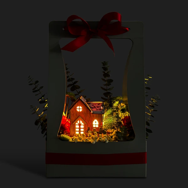 A miniature Christmas village scene with a lit house, pine trees, and red ribbon in a decorative box.