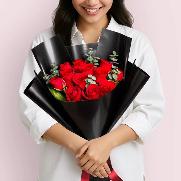 A bouquet of 12 red roses with eucalyptus, wrapped in black paper, held by a person.