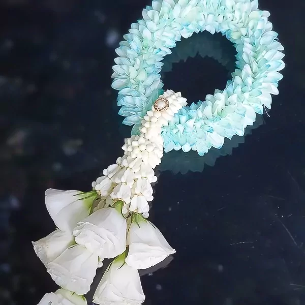 An intricate traditional flower garland lies diagonally on a dark, reflective surface. The upper part of the garland is a thick circular loop crafted from densely arranged light blue and pale green petals. Where the loop connects, a small white woven section is adorned with a round gold and pearl ornament. Cascading downwards from this point is a tightly woven strand of small white flower buds, which finishes with a clustered bouquet of several large, pristine white rosebuds with fresh green sepals.