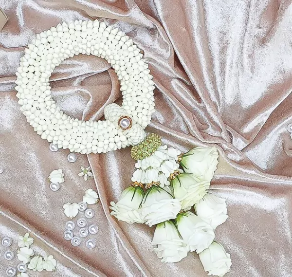An intricate, traditional white flower garland rests diagonally on a draped, shimmering rose gold velvet fabric. The top section forms a thick, circular ring woven tightly with small, white jasmine-like flower buds, secured with a golden and pearl accent where the loop connects. Hanging from the bottom of the loop is a decorative green textured element that transitions into several cascading strands, each ending in a large, partially opened white rose with soft green sepals. A few loose white petals and scattered white pearls are arranged neatly around the garland on the fabric.