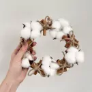 Cotton flower and cinnamon stick wreath with twine bows held by a hand.