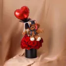 Graduation teddy bear wearing cap, holding heart balloon, surrounded by red roses in a black box.