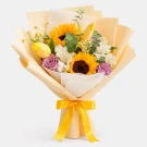 Bouquet of sunflowers, purple roses, white flowers, and yellow tulips wrapped in yellow paper with a yellow bow.