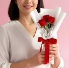 A woman in a white shirt holds a bouquet of three red roses wrapped in white paper and tied with a red ribbon.
