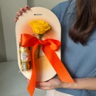 A yellow rose and Ferrero Rocher chocolate tied with an orange ribbon on a light pink oval card.