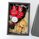 A gift box with red soap roses, a teddy bear keychain, and fairy lights on straw filler.