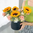 A black box brimming with sunflowers, pink roses, and white carnations, held by a person in a green top.
