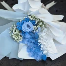 Blue dried rose, hydrangea, and accent flowers in a serene bouquet wrapped in white and blue paper.