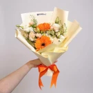 Hand holds a bouquet of orange gerbera daisies, white roses, and delicate green foliage, tied with an orange ribbon.
