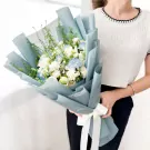 A person holds a bouquet of white and light blue flowers wrapped in pale blue paper with a white ribbon.