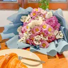 Pink and purple mums with white statice in blue wrapping next to a croissant and fork.