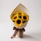 Bouquet of five bright sunflowers wrapped in brown and cream paper, tied with a brown ribbon, on a light background.
