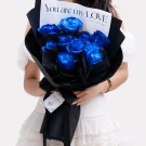 A bouquet of vibrant blue roses wrapped in black paper, held by a person in a white dress.
