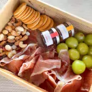 Charcuterie box with Serrano ham, crackers, nuts, fruit, and two small jars of jam and honey.