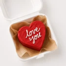 Red heart-shaped cake with "love you" written in white icing, inside a white box lined with brown paper.