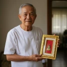 Elderly man in white shirt holds framed Emerald Buddha with gold frame and red backing.