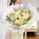 Woman holding a large bouquet of white roses, gerberas, daisies, and yellow craspedias wrapped in white paper.