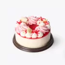 Round strawberry yogurt cake topped with pink and white frosting, white chocolate, and red sprinkles on a dark base.